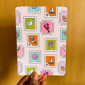 Pode incluir: Um cartão retangular rosa com um padrão repetido de desenhos coloridos de selos postais. Os selos retratam várias imagens, incluindo uma concha, uma lagosta, um veleiro e flores. O cartão é segurado por uma mão.