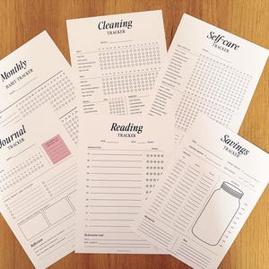 May include: A collection of six white paper tracking sheets with black text and graphics. The sheets include a Monthly Habit Tracker, Cleaning Tracker, Self-care Tracker, Journal Tracker, Reading Tracker, and Savings Tracker with a jar illustration.