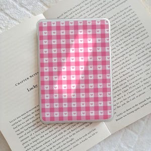 Può includere: Una custodia per telefono rosa e bianca a quadretti con piccoli cuori rosa. La custodia è appoggiata su un libro aperto con testo.