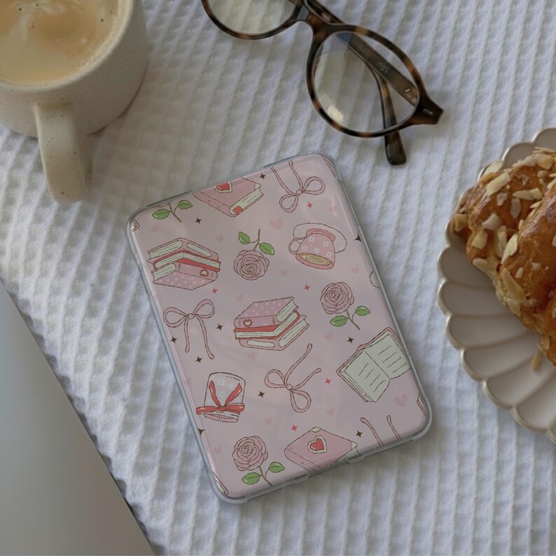 May include: A clear phone case with a pink background and a pattern of books, roses, bows, and teacups. A cup of coffee, a pair of glasses, and a croissant are also visible on a white textured surface.