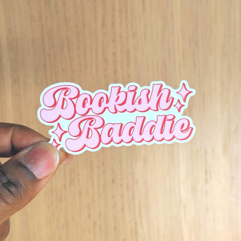 May include: A pink and white sticker with the words "Bookish Baddie" in a retro font. The sticker has a light blue background and star accents. The sticker is held by a person's hand.