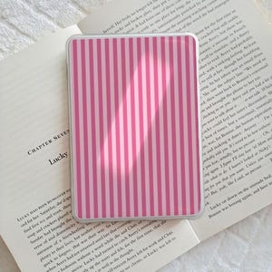 Può includere: Custodia per telefono a righe rosa e bianche. La custodia presenta un motivo a righe verticali e una finitura lucida. È appoggiata su un libro aperto con testo.