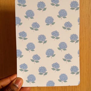 Peut inclure: Une carte rectangulaire aux coins arrondis avec un motif répété de fleurs d'hortensia bleues avec des feuilles vertes sur un fond crème. La carte est tenue par une main sur une surface en bois.