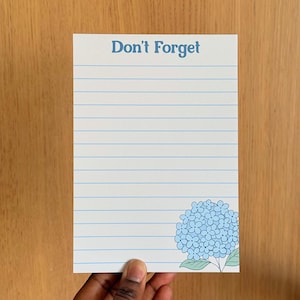 Peut inclure: Bloc-notes blanc avec des lignes horizontales bleues et l'inscription "Don't Forget" en bleu. Une illustration d'hortensia bleu est dans le coin inférieur droit. Le bloc-notes est tenu par une main.