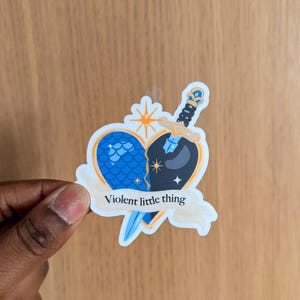 Pode incluir: Um adesivo com um coração dividido em duas metades, uma azul com um padrão de escamas, a outra preta com estrelas. Uma espada perfura o coração, e uma faixa diz "Violent little thing".