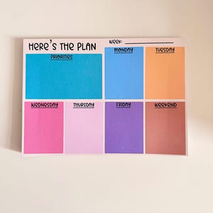 May include: A rectangular weekly planner notepad with the text Here's the Plan and Week at the top. It features a large blue Priorities section and six smaller colored boxes labeled Monday, Tuesday, Wednesday, Thursday, Friday, and Weekend.
