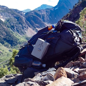 May include: A large, dark blue hiking backpack with a gray box attached, featuring a mountain logo. The backpack is resting on a rocky hillside with a scenic mountain backdrop. A water bottle and other gear are visible.