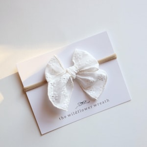 May include: A white embroidered bow on a beige headband, displayed on a white card. The bow features a delicate floral pattern. The card reads "the wildflower wreath" in a script font.