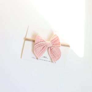 May include: A pale pink baby bow headband. The bow is made of a textured fabric and is attached to a neutral-coloured elastic band. The bow is centered on a white card with the text "the wildflower wren" printed on it.