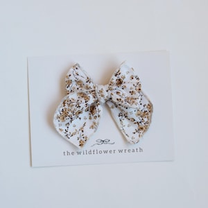 May include: A fabric hair bow featuring a white background with a brown floral pattern. The bow is attached to a white display card that includes a small bow icon and the text "the wildflower wreath" printed in black at the bottom centre.