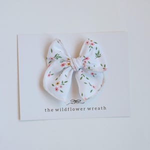 May include: A white fabric hair bow featuring a delicate floral pattern with small pink and yellow flowers and green leaves. The bow is attached to a white display card that includes a small bow icon and the text the wildflower wreath.