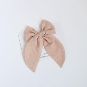 May include: A pale peach hair bow with a polka dot design. The bow is knotted in the middle with long, trailing ends. The bow is made of a soft, textured material and is likely used as a hair accessory.