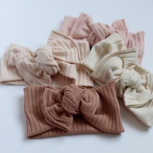 May include: Several ribbed knit headbands in a palette of neutral tones, such as beige, pink, and brown. Each headband has a knotted bow, providing a soft and fashionable accessory.