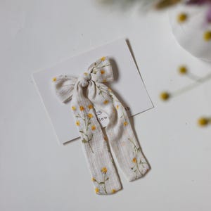 Puede incluir: Un lazo para el cabello hecho de tela color hueso con una textura vertical sutil y un estampado de pequeñas flores amarillas y tallos verdes. El lazo tiene dos extremos largos que cuelgan y está sujeto a una tarjeta rectangular blanca.