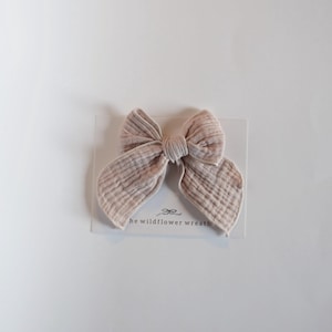 May include: A light pink fabric bow with a textured appearance, resting on a white card. The bow is tied in the center and has two large loops on each side. The card reads "the wildflower wreath".