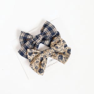 Peut inclure: Deux nœuds pour cheveux en tissu. L'un présente un motif à carreaux bleu marine et beige. L'autre a un fond beige avec un motif floral bleu. Les deux nœuds sont noués au centre et sont posés sur une surface blanche.