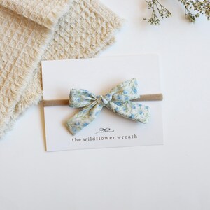 May include: A floral bow in light blue with a beige headband, presented on a white card with the text "the wildflower wreath". The bow has a pattern of small blue flowers and green leaves.