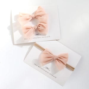 May include: Two light peach-coloured tulle bows attached to white display cards. One card holds two bows, while the other features a single bow on a thin, tan elastic headband. Each card displays the text 'the wildflower wreath' in black font.