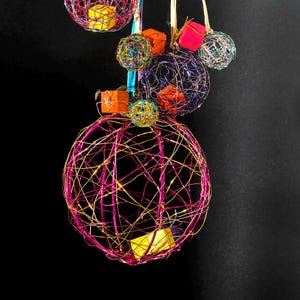 Puede incluir: Una colección de esferas decorativas colgantes hechas de alambre colorido. Las esferas son de varios tamaños y colores, incluyendo rosa, amarillo, morado y dorado. Pequeñas cajas de regalo coloridas se colocan dentro de algunas de las esferas. Las esferas cuelgan de cintas coloridas.