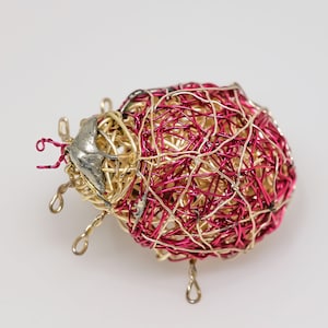 May include: A wire sculpture of a ladybird with a red and gold body. The sculpture is made of thin wire that is twisted and woven together to create a detailed and intricate design.