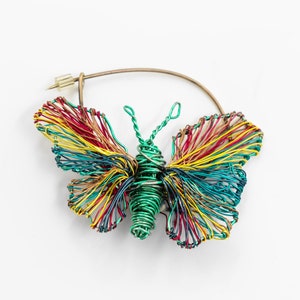 Könnte beinhalten: Eine farbige Draht-Schmetterlingsbrosche mit grünem Körper und roten, gelben und blauen Flügeln. Die Brosche ist an einem goldenen Drahtring befestigt.