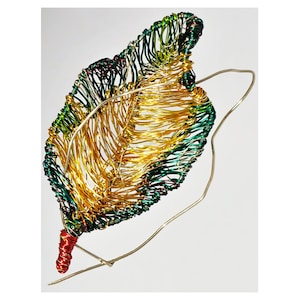 Pode incluir: Uma escultura de folha de arame dourado, verde e vermelho. A folha é feita de arame que foi torcido e tecido para criar uma forma de folha realista. A escultura está suspensa em um fino laço de arame.
