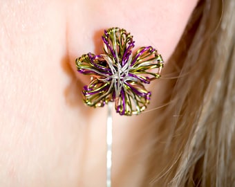 May include: A flower-shaped earring crafted from intertwined metallic wire in shades of purple, olive green, and silver. The five-petaled design features a central cluster of silver wire, with a single straight silver wire stem extending downwards.