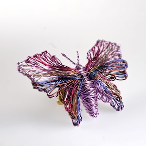 May include: A butterfly-shaped sculpture crafted from thin, iridescent metallic wire. The wings feature a complex, open-work pattern in shades of purple, magenta, blue, and copper, while the central body is tightly coiled with purple wire.