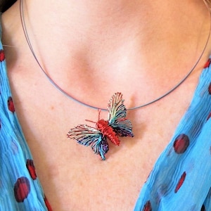 Puede incluir: Un delicado collar con colgante de mariposa. La mariposa está hecha de alambre rojo, azul y verde, suspendida de un collar azul fino. El fondo presenta una bufanda azul con lunares rojos.