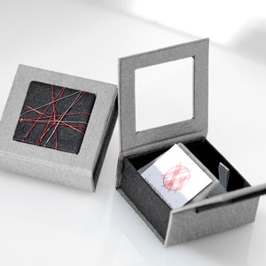 May include: Two square gray gift boxes. One box is closed, featuring a black square top decorated with crisscrossing red and silver threads. The second box is open, revealing a black interior and a white card with a red circular graphic design.