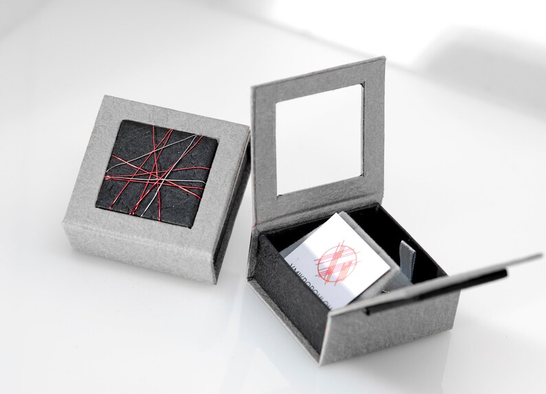 May include: Two square gray gift boxes. One box is closed, featuring a black square top decorated with crisscrossing red and silver threads. The second box is open, revealing a black interior and a white card with a red circular graphic design.