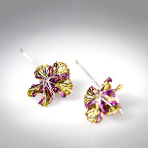 May include: A pair of dangle earrings crafted from thin, intertwined metallic wire in vibrant shades of purple, lime green, and silver. The wire is shaped into a multi-petaled flower design, with each earring featuring a simple silver-toned hook.