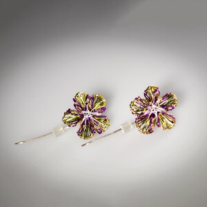 May include: A pair of stud earrings featuring five-petaled flower shapes crafted from thin, intertwined metallic wire in shades of vibrant purple and lime green, with a central silver-toned wire detail and clear plastic earring backings.