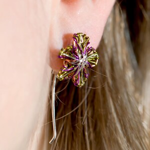 May include: A wire-wrapped earring featuring a multi-petaled flower design crafted from metallic purple, lime green, and silver-toned wire. The floral element is attached to a long, slender, smooth silver-toned metal post that extends downward.