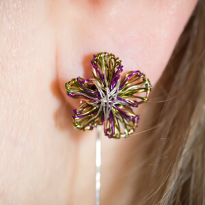 Puede incluir: Un par de pendientes con un diseño de flor hecho de alambre. La flor es de color morado, verde y plateado. Los pendientes cuelgan de la oreja de una persona.