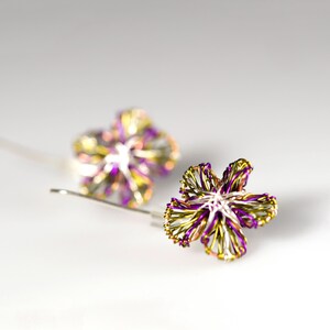 May include: A pair of earrings featuring flower-shaped designs crafted from thin, intertwined metallic wire in shades of purple, lime green, and silver. Each earring includes a simple hook wire for attachment, showcasing a delicate, open-work structure.