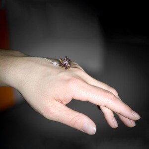 May include: A ring featuring a multi-colored, iridescent flower-shaped centerpiece with purple, green, and gold tones. The flower is attached to a thin, silver-toned metal band that rests on the back of the hand, showcasing a delicate design.