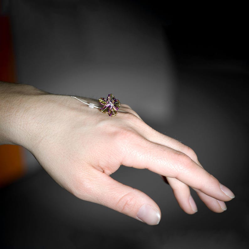 May include: A ring featuring a multi-colored, iridescent flower-shaped centerpiece with purple, green, and gold tones. The flower is attached to a thin, silver-toned metal band that rests on the back of the hand, showcasing a delicate design.