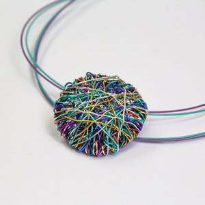 Puede incluir: Un collar de alambre colorido con un colgante redondo hecho de alambres entrelazados en tonos de morado, azul, verde, dorado y plateado.