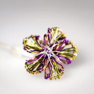 May include: A decorative hair pin featuring a flower shape constructed from intertwined metallic wires in shades of purple, lime green, copper, and silver. The wire petals are looped and layered to create a multi-dimensional floral design.