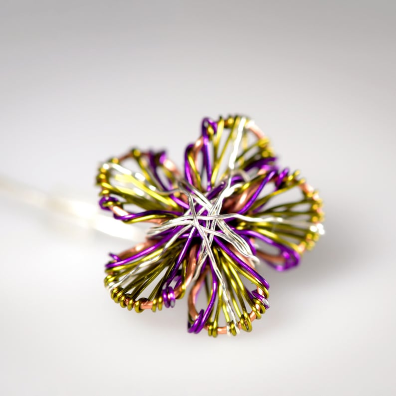 May include: A decorative hair pin featuring a flower shape constructed from intertwined metallic wires in shades of purple, lime green, copper, and silver. The wire petals are looped and layered to create a multi-dimensional floral design.