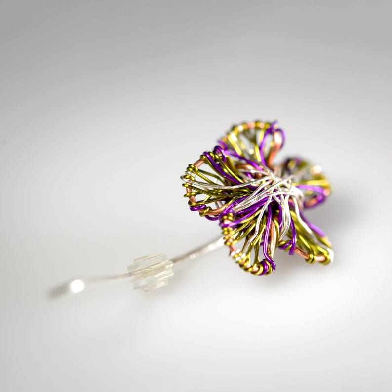 May include: A flower-shaped brooch crafted from intertwined metallic wires in shades of purple, lime green, and copper. The center features a dense cluster of silver wire, and a clear plastic clutch back is attached to the pin stem on the side.