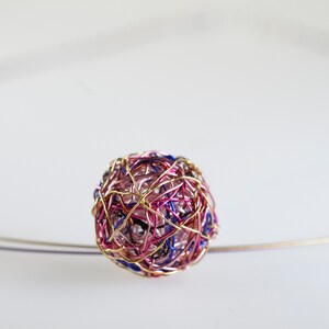 Op de afbeelding: Een roze, blauwe en gouden draadbol ornament. De bol is gemaakt van dunne draad die is gedraaid en geweven om een uniek en ingewikkeld ontwerp te creëren.