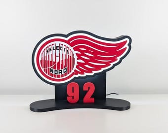 May include: A black and red illuminated sign featuring a stylized wing design and the number 92. The sign includes the text "SELECTS NORD" within a circular emblem. The design is set on a black base.