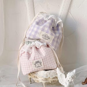 Pode incluir: Duas sacolas com cordão xadrez, uma rosa e outra lavanda. Cada sacola tem uma borda de renda branca e uma etiqueta que diz "HAND MADE" com detalhes florais. As sacolas estão em um suporte decorativo, junto com uma pequena estátua de anjo.