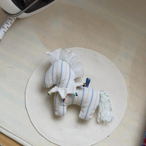 May include: A handmade stuffed horse toy with blue striped fabric and a white mane. The horse has a white collar with colorful beads and a blue ribbon. It is placed on a white circular surface.