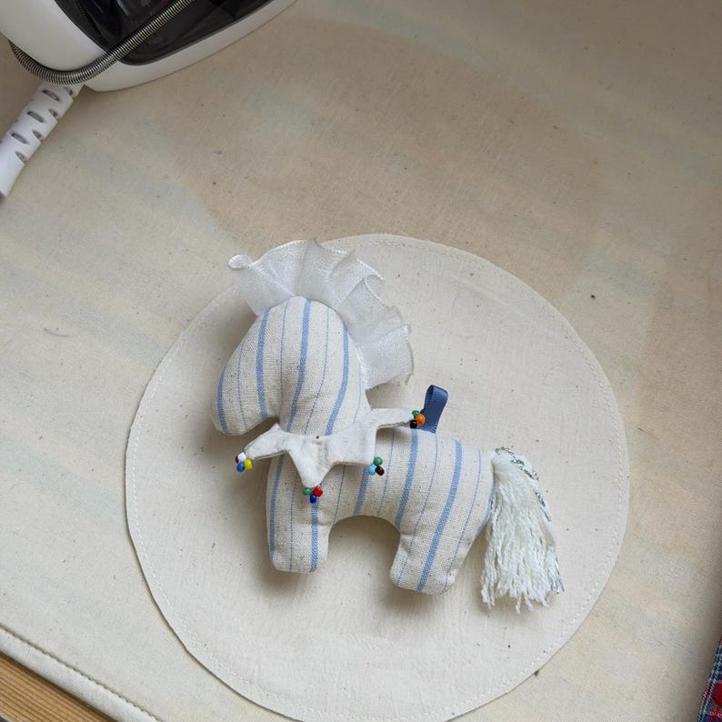 May include: A handmade stuffed horse toy with blue striped fabric and a white mane. The horse has a white collar with colorful beads and a blue ribbon. It is placed on a white circular surface.