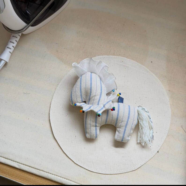 May include: A handmade stuffed horse toy with blue and white striped fabric. The horse has a white mane and tail, and colorful pins are used to hold the fabric together. The toy is on a circular piece of fabric.