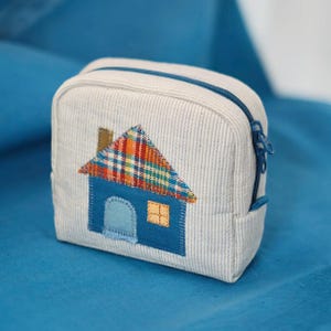 Puede incluir: Pequeño neceser cuadrado de tela con estampado de rayas azules y blancas. Una aplicación de casa azul con tejado a cuadros y ventana naranja está centrada en la parte delantera. El neceser tiene una cremallera azul y un pequeño lazo lateral.