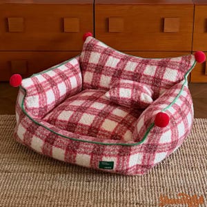 Può includere: Un'accogliente cuccia per animali domestici con un motivo a quadri rossi e bianchi. Il letto ha una forma arrotondata con un piccolo cuscino ed è rifinito con bordini verdi. I pompon rossi adornano gli angoli. Il letto è realizzato in tessuto morbido e strutturato.